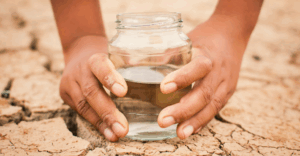 Two hands holding a glass jar filled with water above cracked, dry earth, symbolizing water scarcity and environmental concerns.